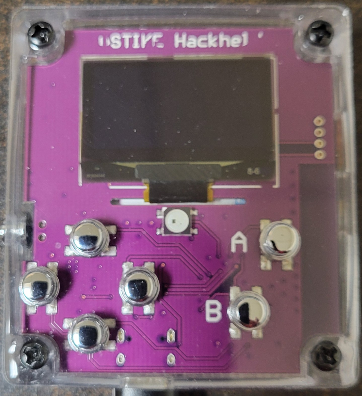 Jeff's DSTIKE Hackheld V1, front view — purple PCB, "DSTIKE Hackheld" silkscreen banner at the top, 0.96″ OLED, and the seven-button cluster (4-way diamond + A/B + reset). The clear acrylic case sh…
